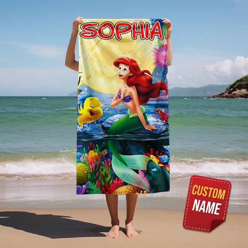 Puede incluir: Una toalla de playa rectangular con un gr&aacute;fico colorido de la sirena Ariel, junto con Flounder y Sebasti&aacute;n. El nombre SOPHIA est&aacute; impreso en letras rojas grandes en la parte superior. El dise&ntilde;o incluye una escena submarina detallada.