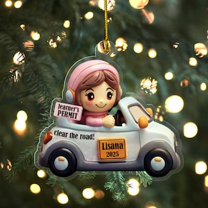 May include: A clear, car-shaped ornament featuring a cartoon character wearing a pink hat and headphones, driving a white car. The ornament has the text "learner's PERMIT", "clear the road!", and "Lisana 2025".