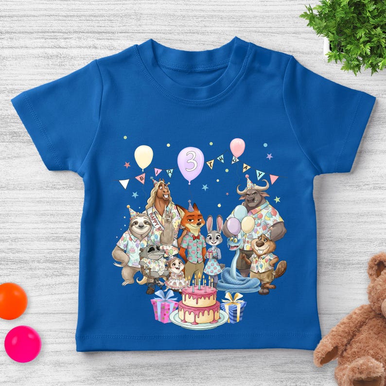 May include: A blue t-shirt featuring a colorful graphic of animated animal characters from a movie, celebrating a birthday. The number "3" is on a balloon. A birthday cake and presents are also shown.