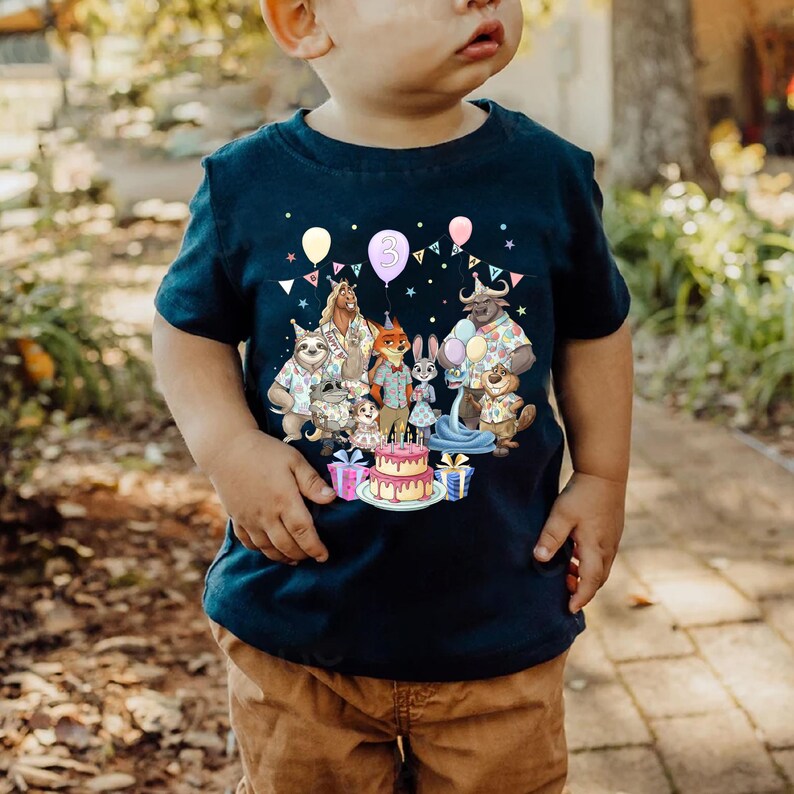 May include: Navy blue toddler t-shirt featuring a colorful graphic of cartoon animals, balloons, and a birthday cake. The number "3" is on a balloon. The shirt is worn by a young child.