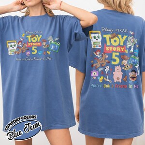 May include: Blue jean-colored t-shirt featuring a graphic from Disney Pixar's Toy Story 5. The front of the shirt displays the Toy Story 5 logo and characters, with the text "You're Got a Friend In Me". The back of the shirt also features the Toy Story 5 graphic.