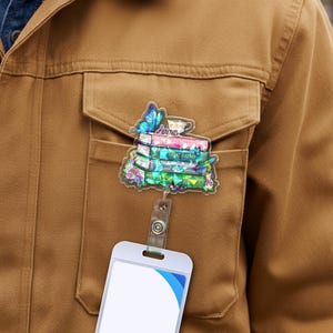 May include: A badge reel featuring a stack of books with the text "The Great Gatsby" and a teacup, a blue butterfly, and the name "Anna". The badge reel is attached to a white card holder.