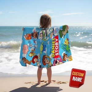 May include: A light blue beach towel featuring a colorful print of various Toy Story characters, including Woody, Buzz Lightyear, and Jessie, with the text DANIEL STORY printed in yellow and white letters against a red background.