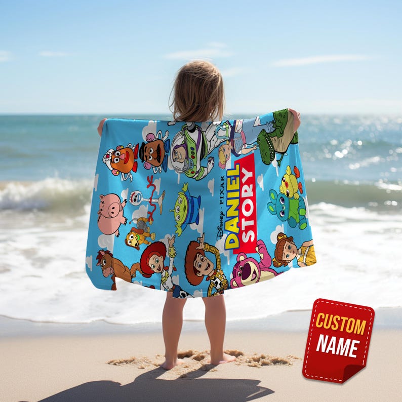 May include: A light blue beach towel featuring a colorful print of various Toy Story characters, including Woody, Buzz Lightyear, and Jessie, with the text DANIEL STORY printed in yellow and white letters against a red background.