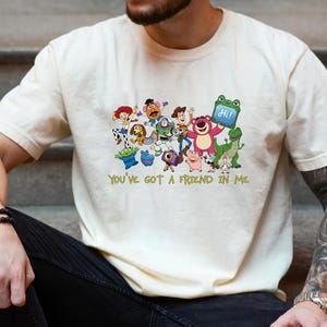 May include: A cream-colored short-sleeve t-shirt featuring a colorful graphic print of various Toy Story characters including Woody, Buzz Lightyear, and Lotso. Below the characters, the text "YOU'VE GOT A FRIEND IN ME" is printed in yellow.