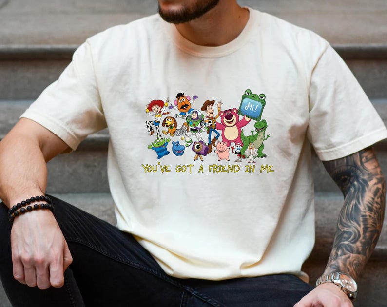 May include: A cream-colored short-sleeve t-shirt featuring a colorful graphic print of various Toy Story characters including Woody, Buzz Lightyear, and Lotso. Below the characters, the text "YOU'VE GOT A FRIEND IN ME" is printed in yellow.