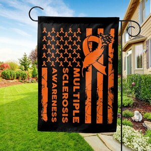 May include: Black garden flag with orange ribbon and text for Multiple Sclerosis Awareness. The flag features an orange ribbon design and the words "Multiple Sclerosis Awareness" in orange. The flag is hanging outside on a metal stand.