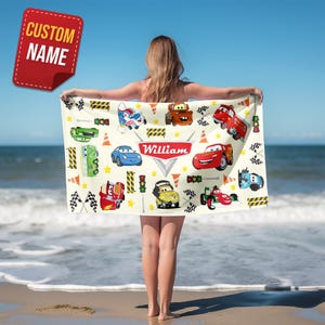 May include: A rectangular beach towel featuring a cream background with a repeating pattern of cartoon race cars, traffic lights, checkered flags, and orange traffic cones. The center displays a red emblem with the name William in white text.