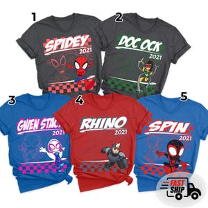 May include: Five colorful t-shirts featuring superhero-themed graphics. The shirts are in various colors: charcoal, dark grey, blue, red, and royal blue. Each shirt has a different character name and the year 2021 printed on it. The shirts are for sale.