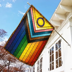 May include: A colorful flag with a stained-glass design, featuring a rainbow gradient and geometric patterns. The flag is mounted on a white house, with a clear blue sky in the background. The flag has a yellow circle with a purple outline.