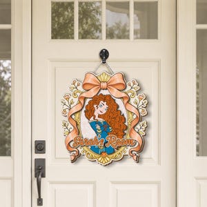 May include: A decorative wooden door sign featuring a cartoon illustration of a girl with long curly red hair wearing a blue dress. The sign is framed by a gold border and a large peach-colored bow, with the text Sarah's Room written in orange.