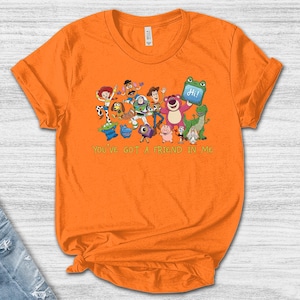 May include: A bright orange short-sleeved t-shirt featuring a colorful graphic print of various animated toy characters and the text "YOU'VE GOT A FRIEND IN ME" printed in green below the characters. The shirt has a crew neckline and a standard fit.