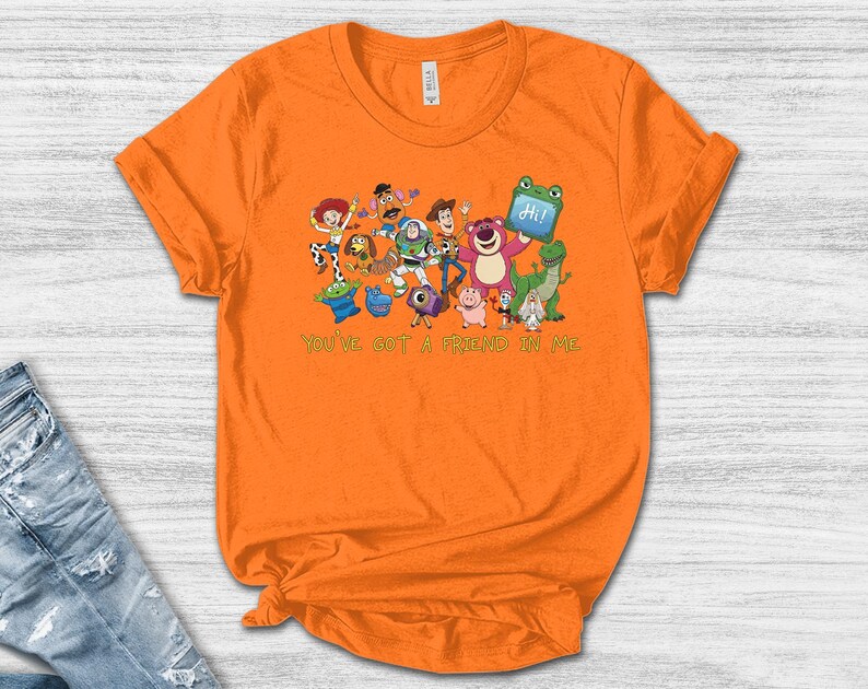 May include: A bright orange short-sleeved t-shirt featuring a colorful graphic print of various animated toy characters and the text "YOU'VE GOT A FRIEND IN ME" printed in green below the characters. The shirt has a crew neckline and a standard fit.