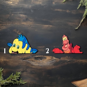 May include: Two colorful wooden cutouts of characters from The Little Mermaid. One is a yellow and blue fish with a smiling face, and the other is a red crab with a surprised expression. The cutouts are placed on a dark wooden surface.