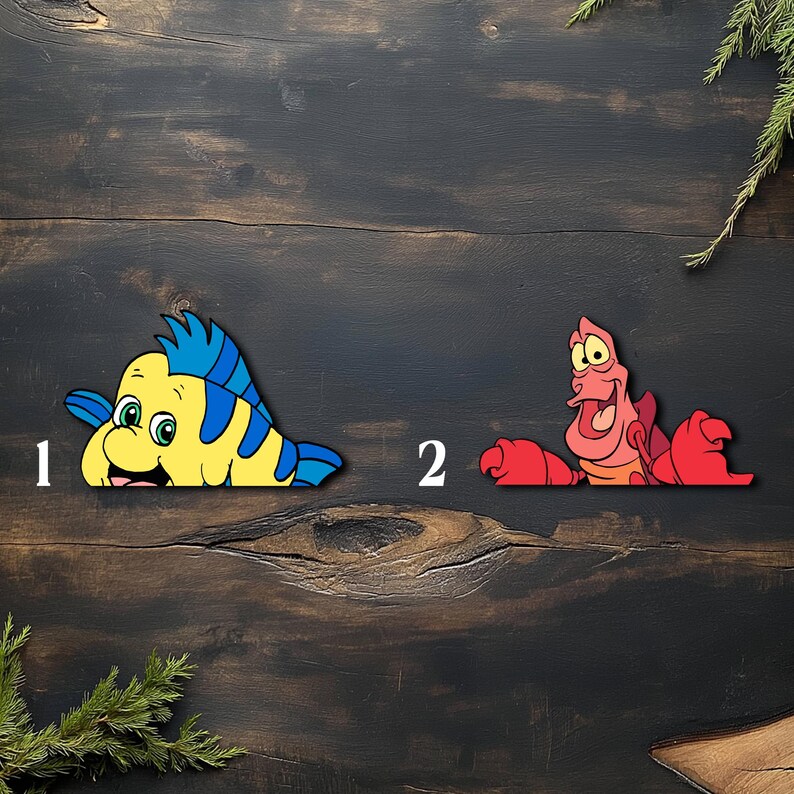 May include: Two colorful wooden cutouts of characters from The Little Mermaid. One is a yellow and blue fish with a smiling face, and the other is a red crab with a surprised expression. The cutouts are placed on a dark wooden surface.