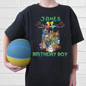 May include: A black t-shirt featuring a colorful graphic of animated animal characters wearing party hats, a birthday cake, and the text "JAMES" at the top and "BIRTHDAY BOY" at the bottom, with a yellow "3" inside a Mickey Mouse icon.