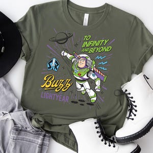May include: An olive green t-shirt featuring a graphic of Buzz Lightyear in a flying pose. The text reads "TO INFINITY AND BEYOND" in green, "Buzz" in yellow script, and "LIGHTYEAR" in purple. The design includes a planet, stars, and a blue emblem.