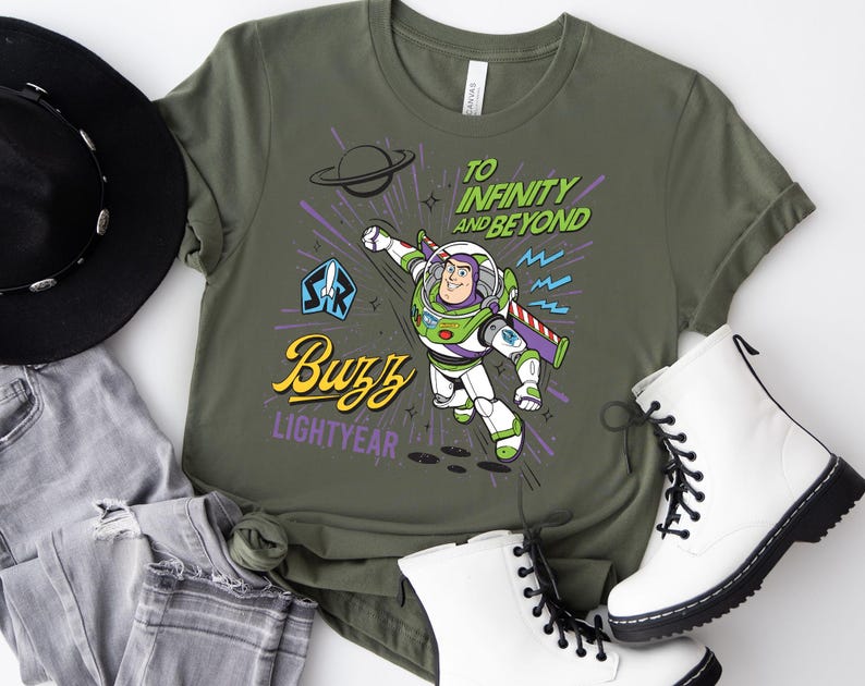 May include: An olive green t-shirt featuring a graphic of Buzz Lightyear in a flying pose. The text reads "TO INFINITY AND BEYOND" in green, "Buzz" in yellow script, and "LIGHTYEAR" in purple. The design includes a planet, stars, and a blue emblem.