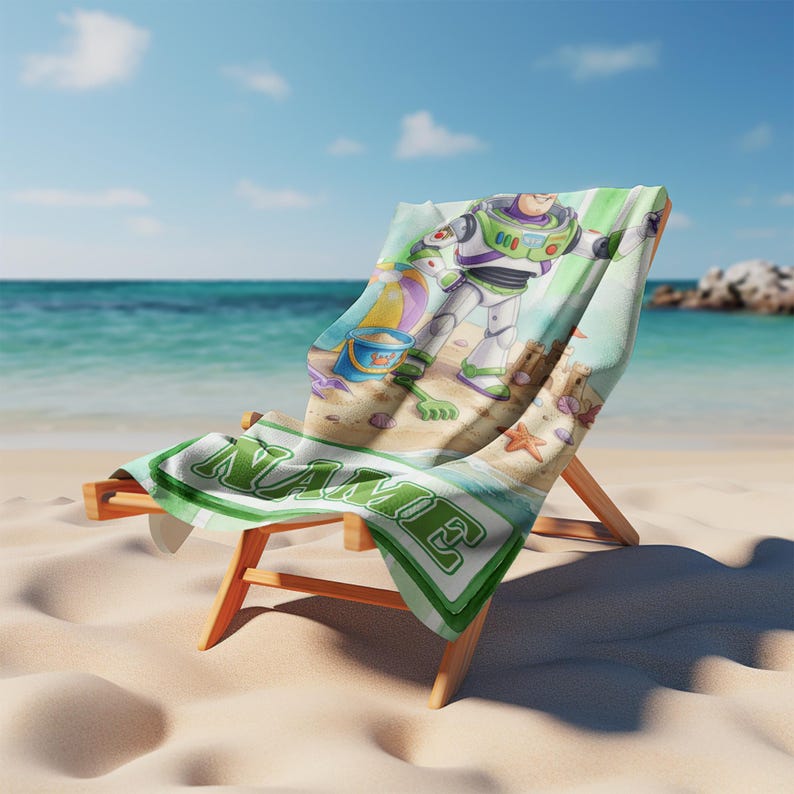May include: A beach towel draped over a wooden lounge chair, featuring a printed illustration of a space ranger character, a beach ball, a blue bucket, and a sandcastle. The towel includes the word NAME in large green block letters.