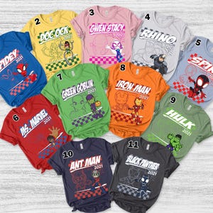 May include: A collection of eleven short-sleeved t-shirts in various colors, each featuring a different superhero graphic and the year 2021. The shirts include characters like Spidey, Ms. Marvel, Doc Ock, Gwen Stacy, Rhino, Spin, Iron Man, Hulk, Ant Man, and Black Panther.