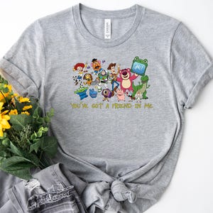 May include: A heather gray short-sleeve t-shirt featuring a colorful graphic print of various animated characters from the Toy Story franchise, including Woody and Buzz Lightyear, with the yellow text "YOU'VE GOT A FRIEND IN ME" printed below.