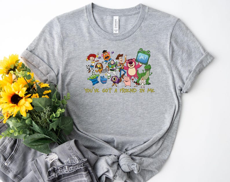 May include: A heather gray short-sleeve t-shirt featuring a colorful graphic print of various animated characters from the Toy Story franchise, including Woody and Buzz Lightyear, with the yellow text "YOU'VE GOT A FRIEND IN ME" printed below.