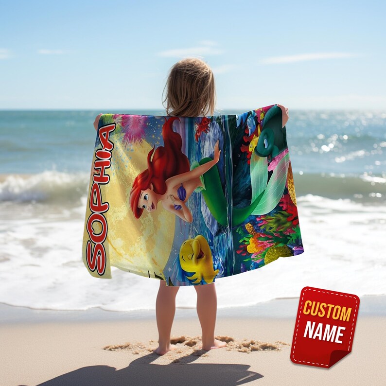 Puede incluir: Una toalla de playa rectangular con una colorida escena submarina que muestra a una sirena de pelo rojo y un pez amarillo. El nombre SOPHIA est&aacute; impreso verticalmente en letras rojas grandes sobre un fondo de luna amarilla.