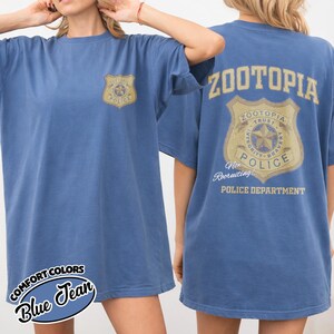 Puede incluir: Camiseta azul vaquero con un gr&aacute;fico de la insignia del Departamento de Polic&iacute;a de Zootopia en la parte delantera y trasera. La parte trasera de la camiseta presenta el texto "ZOOTOPIA" encima de la insignia. La camiseta est&aacute; etiquetada como "COMFORT COLORS" y "Blue Jean".