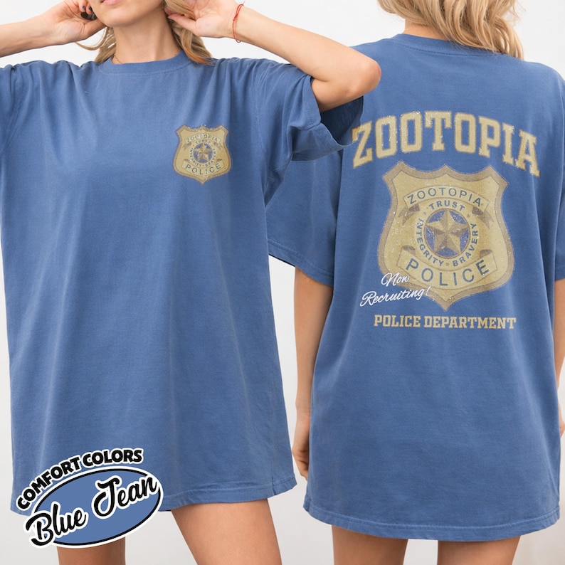Puede incluir: Camiseta azul vaquero con un gr&aacute;fico de la insignia del Departamento de Polic&iacute;a de Zootopia en la parte delantera y trasera. La parte trasera de la camiseta presenta el texto "ZOOTOPIA" encima de la insignia. La camiseta est&aacute; etiquetada como "COMFORT COLORS" y "Blue Jean".