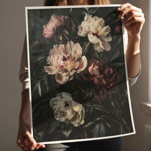 May include: A floral print featuring a dark background with large, detailed peony blooms in shades of pink, cream, and burgundy. The artwork has a vintage aesthetic, with visible brushstrokes and a moody, romantic feel. The print is held by a person.