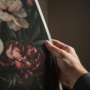 May include: A floral print featuring large peonies in shades of burgundy, cream, and green. The artwork has a dark, moody aesthetic, with the flowers' details and textures prominently displayed. The print is held by a person.