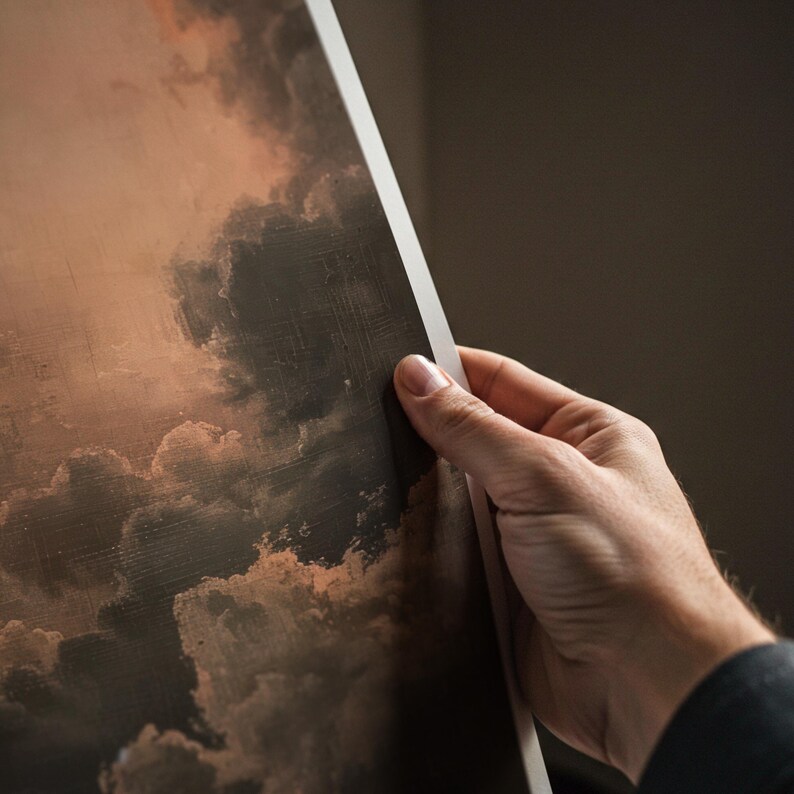 Peut inclure: Une impression d'un paysage nuageux dramatique dans les tons de marron, de gris et de p&ecirc;che. L'&oelig;uvre est tenue par une main, r&eacute;v&eacute;lant une bordure blanche. L'image a un aspect vintage et textur&eacute;.