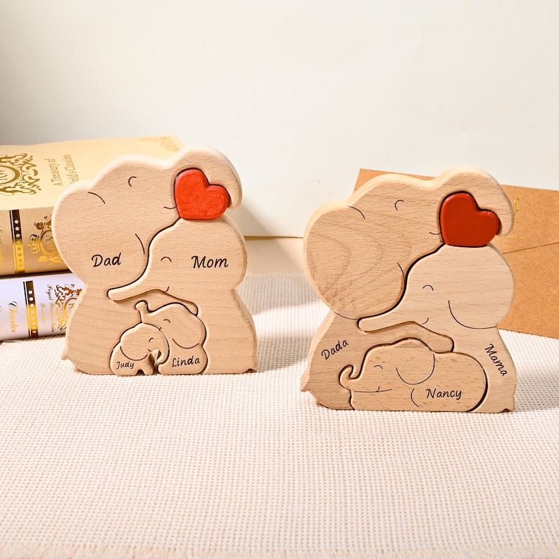 K&ouml;nnte beinhalten: Zwei Holzpuzzles mit Elefantenfamilien. Jedes Puzzle zeigt einen Eltern-Elefanten mit rotem Herz, einen kleineren Elefanten und die Namen "Dad", "Mom", "Judy" und "Linda" auf dem einen und "Dada", "Mama" und "Nancy" auf dem anderen.