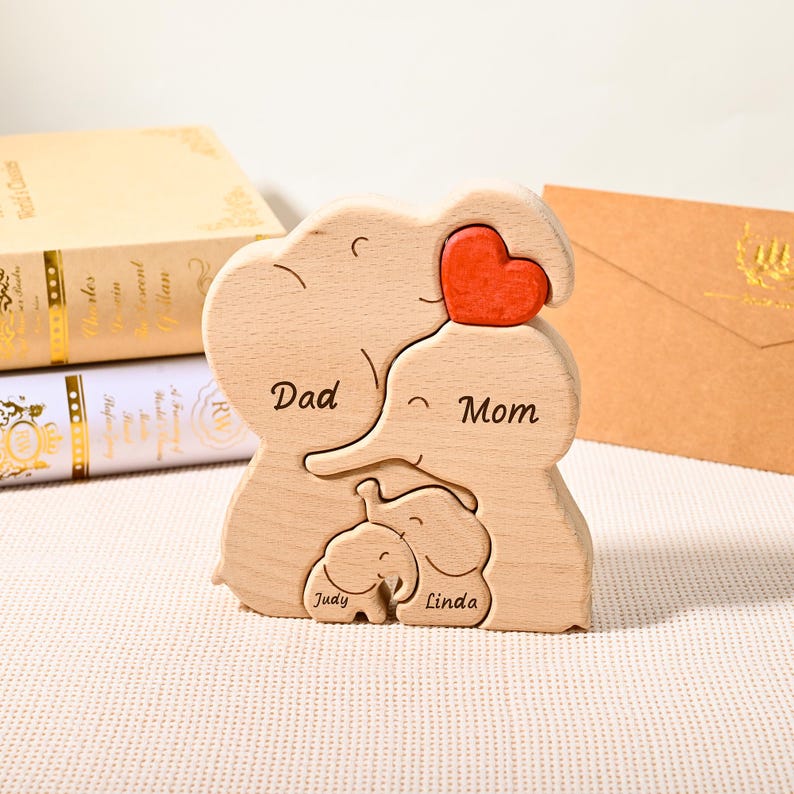 K&ouml;nnte beinhalten: Holzpuzzle f&uuml;r die Familie in Form von Elefanten. Das Puzzle enth&auml;lt die W&ouml;rter "Dad", "Mom", "Judy" und "Linda". Ein rotes Herz ist oben platziert. Das Puzzle ist aus hellem Holz gefertigt.
