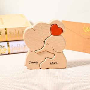 K&ouml;nnte beinhalten: Ein Holzpuzzle mit zwei sich umarmenden Elefanten und einem roten Herz. Die Namen "Jenny" und "Mike" sind auf der Basis eingraviert. Das Puzzle ist aus hellem Holz gefertigt.