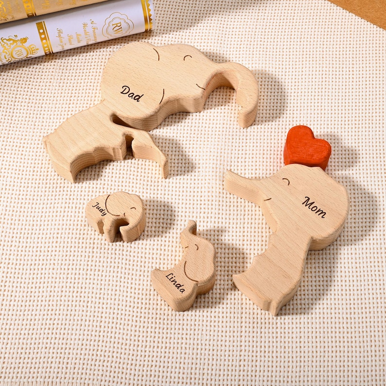 K&ouml;nnte beinhalten: Holz-Elefanten-Familien-Puzzleset. Das Set enth&auml;lt einen gro&szlig;en Elefanten mit der Gravur "Dad", einen kleineren Elefanten mit "Judy", einen weiteren kleinen Elefanten mit "Linda" und einen Elefanten mit Herz und dem Wort "Mom".