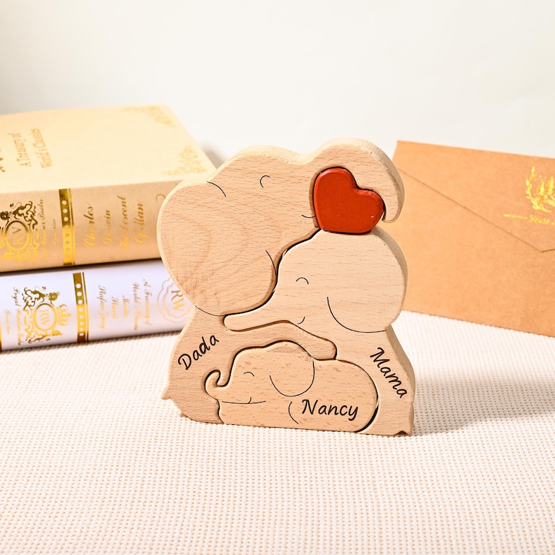 K&ouml;nnte beinhalten: Holz-Elefanten-Familienpuzzle mit rotem Herz. Das Puzzle zeigt drei Elefanten mit den Worten "Dada", "Mama" und "Nancy". Das Puzzle ist aus hellem Holz gefertigt.