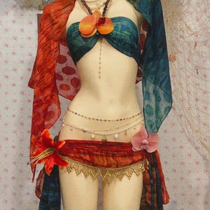 以下が含まれることがあります： マネキンに展示されたベリーダンスの衣装。 このアンサンブルには、ティールとオレンジの模様のトップス、お揃いのショール、赤と金のヒップスカーフが含まれています。 この衣装は、ビーズのネックレス、ウエストチェーン、花のアクセントで飾られています。