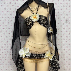 Puede incluir: Un disfraz de danza del vientre con estampado floral en blanco y negro con un velo a juego. El disfraz incluye un top halter, una falda y accesorios con cuentas. Acentos de flores blancas y amarillas están unidos al disfraz.