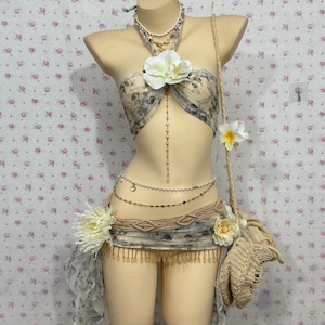 Puede incluir: Un maniquí exhibe un atuendo de estilo bohemio. El conjunto incluye un top halter estampado, una falda con flecos y un cinturón con cuentas. Los accesorios incluyen un bolso tejido, collares y detalles florales. La paleta de colores es neutra con detalles florales y de encaje.