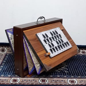 May include: A wooden harmonium with a silver handle and a keyboard of black keys. The instrument has a wooden frame with a hinged top and sides. The interior features decorative panels with gold and purple accents.
