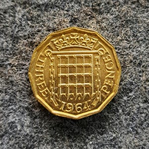 Pode incluir: Uma moeda dourada, octogonal, de três pence de 1964. A moeda apresenta um padrão de grade, uma coroa e as palavras "THREE PENCE" na borda. A moeda está sobre um fundo cinza texturizado.