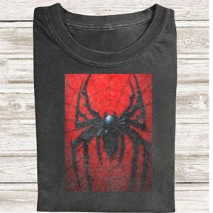 May include: Dark grey t-shirt with a striking spider design. The graphic shows a black spider against a red web backdrop. The t-shirt is folded, suggesting a comfortable fabric.