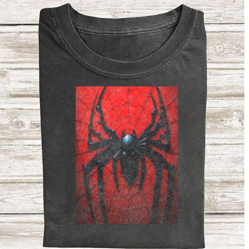 May include: Dark grey t-shirt with a striking spider design. The graphic shows a black spider against a red web backdrop. The t-shirt is folded, suggesting a comfortable fabric.
