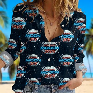 Op de afbeelding: Zwart overhemd met een herhalend patroon van een planeet met een raket, sterren en de tekst "COPY MOON JOY" in wit, blauw en roze. Het shirt met lange mouwen heeft opgerolde manchetten.