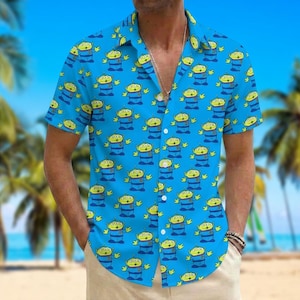Op de afbeelding: Een felblauw overhemd met korte mouwen en een knoopsluiting, voorzien van een herhalend patroon van groene drieogige buitenaardse wezens in blauwe uniformen. Het overhemd heeft een klassieke kraag en een verticale rij witte knopen aan de voorzijde.