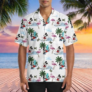 Puede incluir: Camisa blanca de manga corta con un estampado repetido de Mickey Mouse surfeando, flamencos rosas, palmeras y tortugas. La camisa la lleva una persona y está ambientada en una escena de playa tropical.