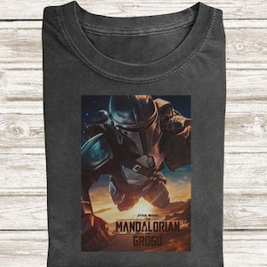May include: Dark grey t-shirt with a graphic from Star Wars: The Mandalorian and Grogu. The image shows the Mandalorian in flight, with a space background. The text "STAR WARS THE MANDALORIAN AND GROGU" is printed at the bottom.