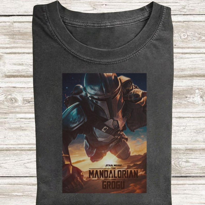 May include: Dark grey t-shirt with a graphic from Star Wars: The Mandalorian and Grogu. The image shows the Mandalorian in flight, with a space background. The text "STAR WARS THE MANDALORIAN AND GROGU" is printed at the bottom.