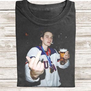 May include: Black t-shirt featuring a photo of a person wearing a USA jersey, holding a beer and cigarette, and giving the middle finger. The jersey has the word "USA" on the front and a blue and red collar.